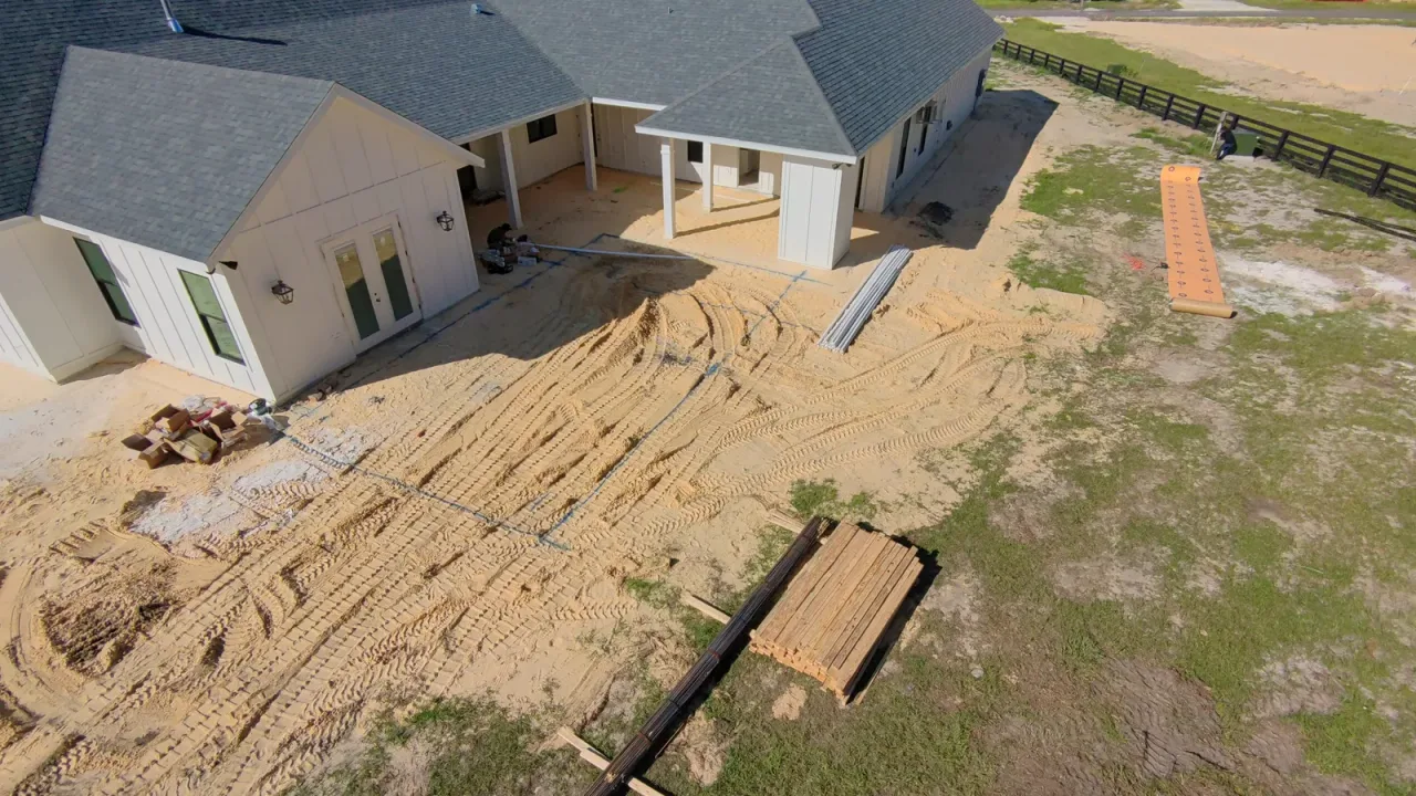 Aerial view of a sandy backyard behind a white house before excavation, with lumber staged and a fenced pasture beyond.