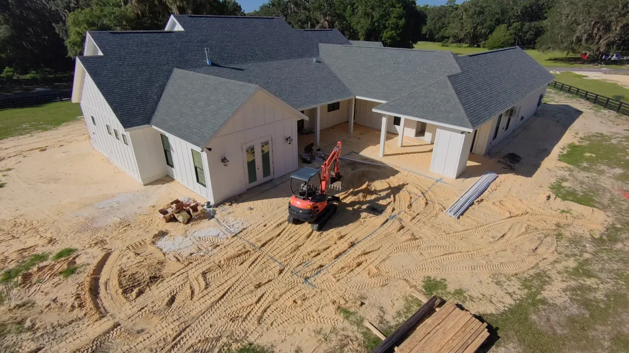 Aerial view of an orange mini excavator parked on the sandy yard behind a white house, ready to dig the pool.