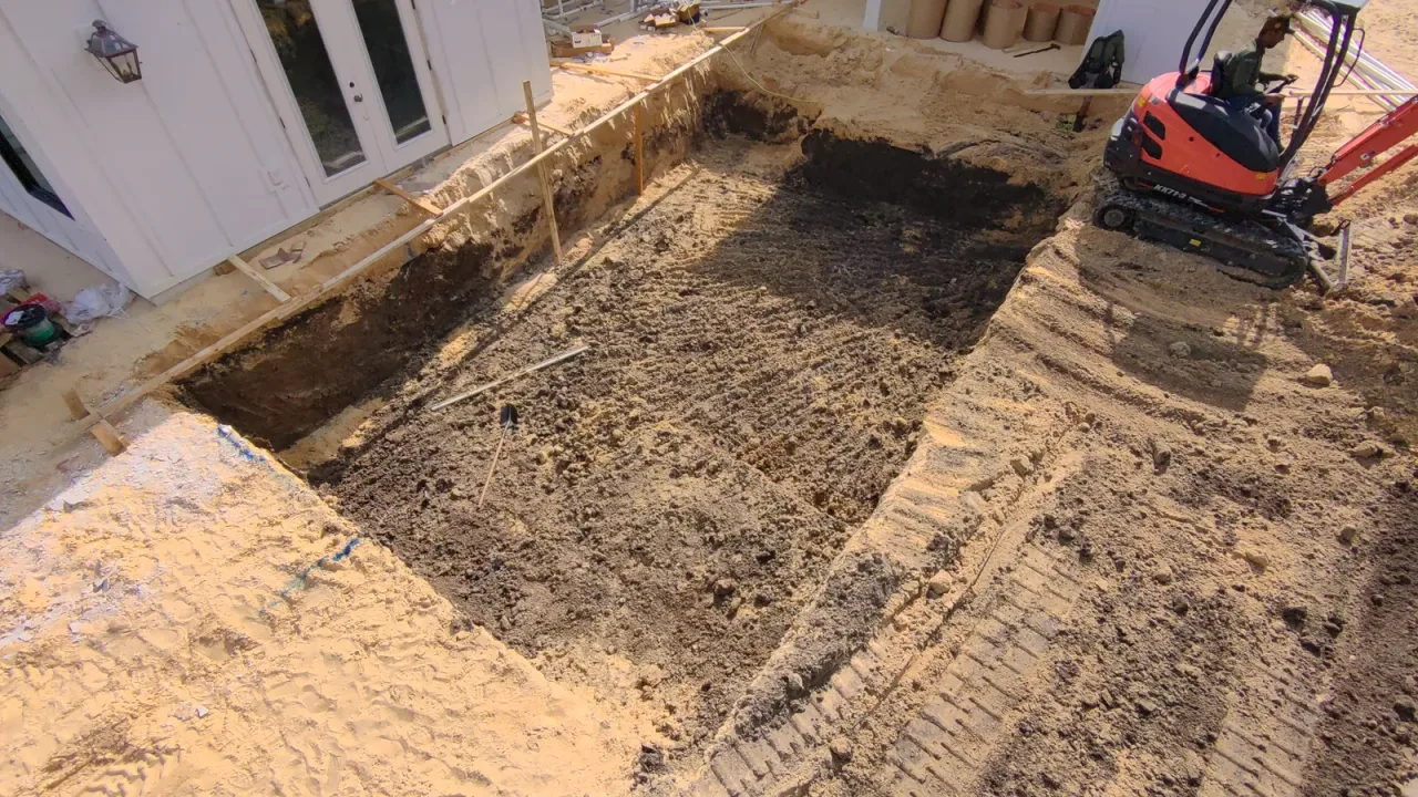 Close aerial view of a dug pool showing its depth, with form boards along the house and a mini excavator at the edge.