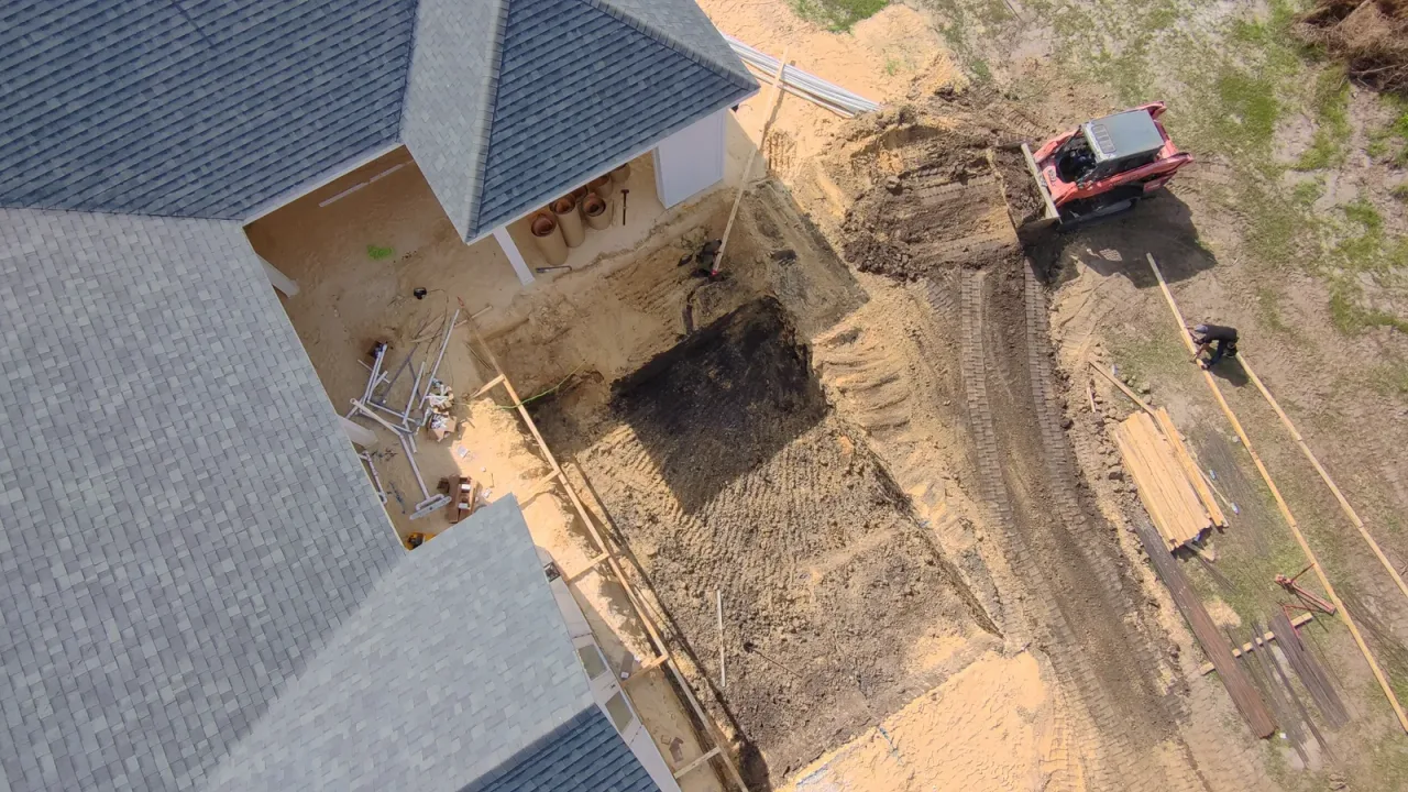 Top-down drone view of a pool excavation beside the house as a skid steer hauls soil away from the dig.