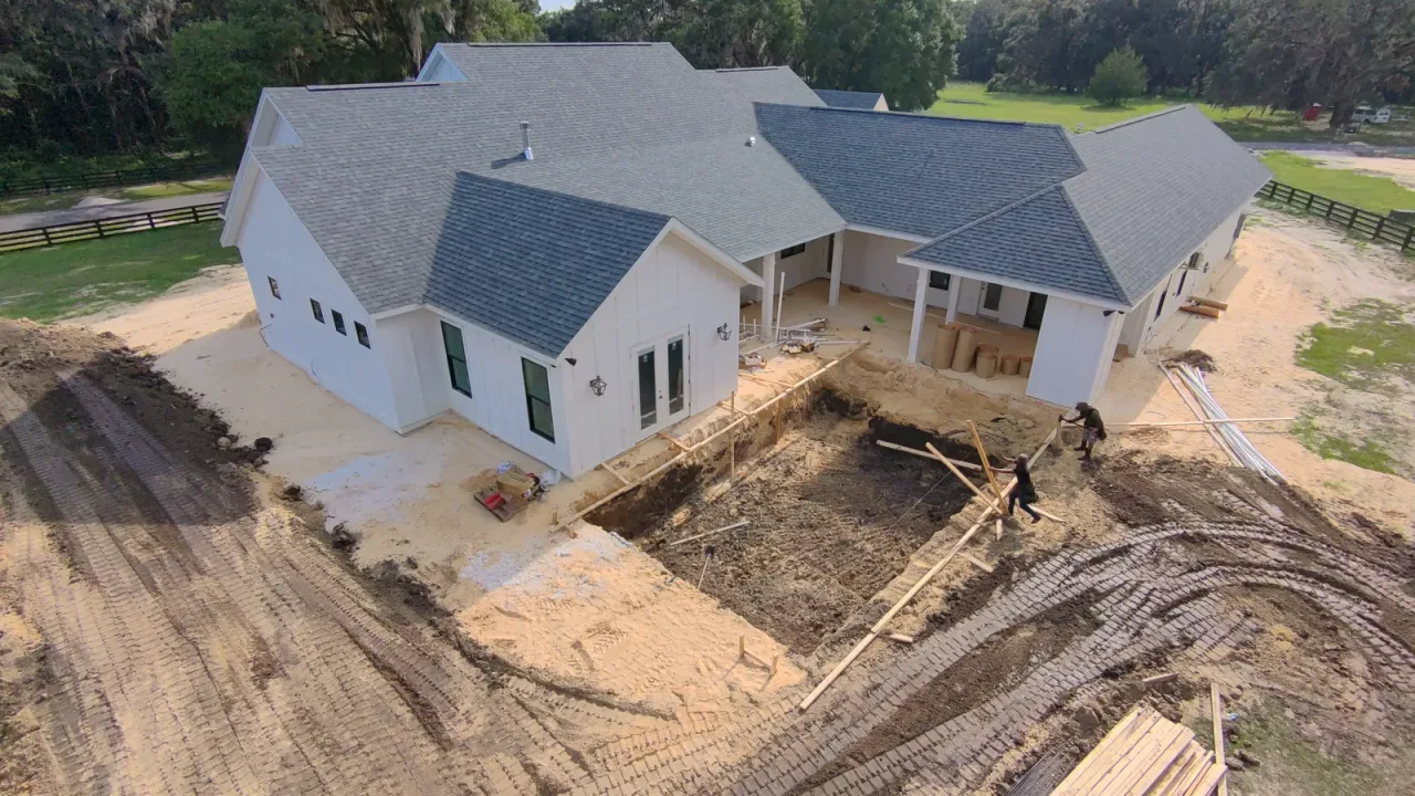 Aerial view of a freshly dug pool hole behind a white farmhouse, with two workers setting wooden form boards.