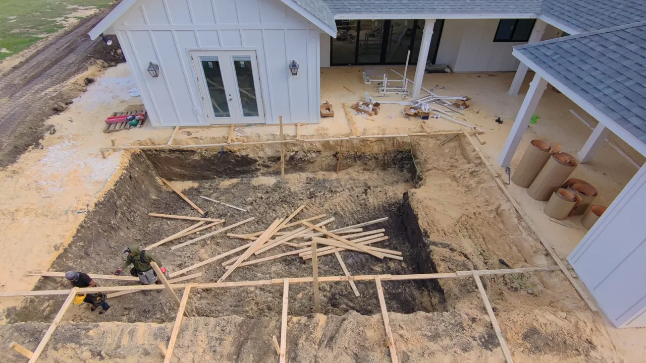 Two workers setting form boards inside a finished pool excavation behind a white house, with lumber laid across the hole.