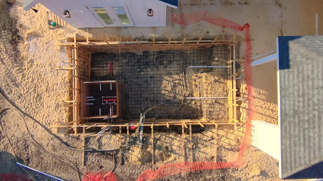 Top-down view of a pool rebar cage in wood forms, with a square spa form, plumbing stubs and orange safety fencing around.
