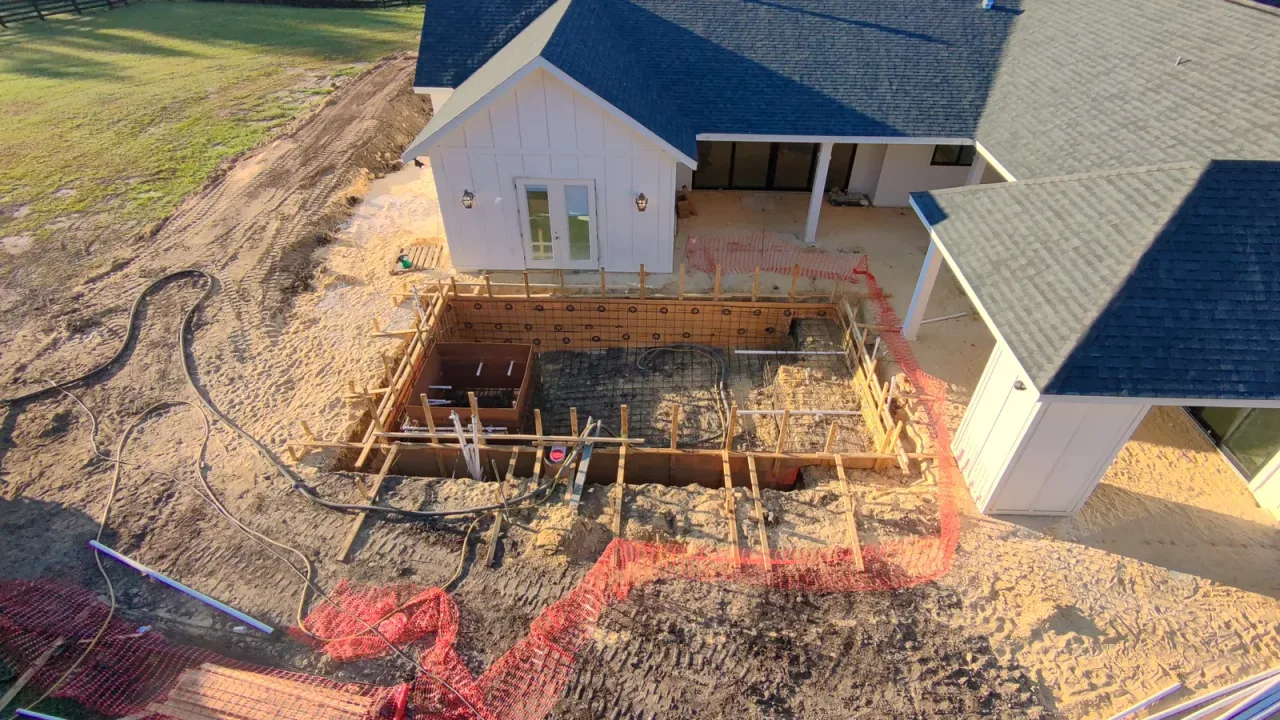 Aerial view of a pool rebar cage and wood forms behind a white house, with orange safety fencing and hoses around the site.