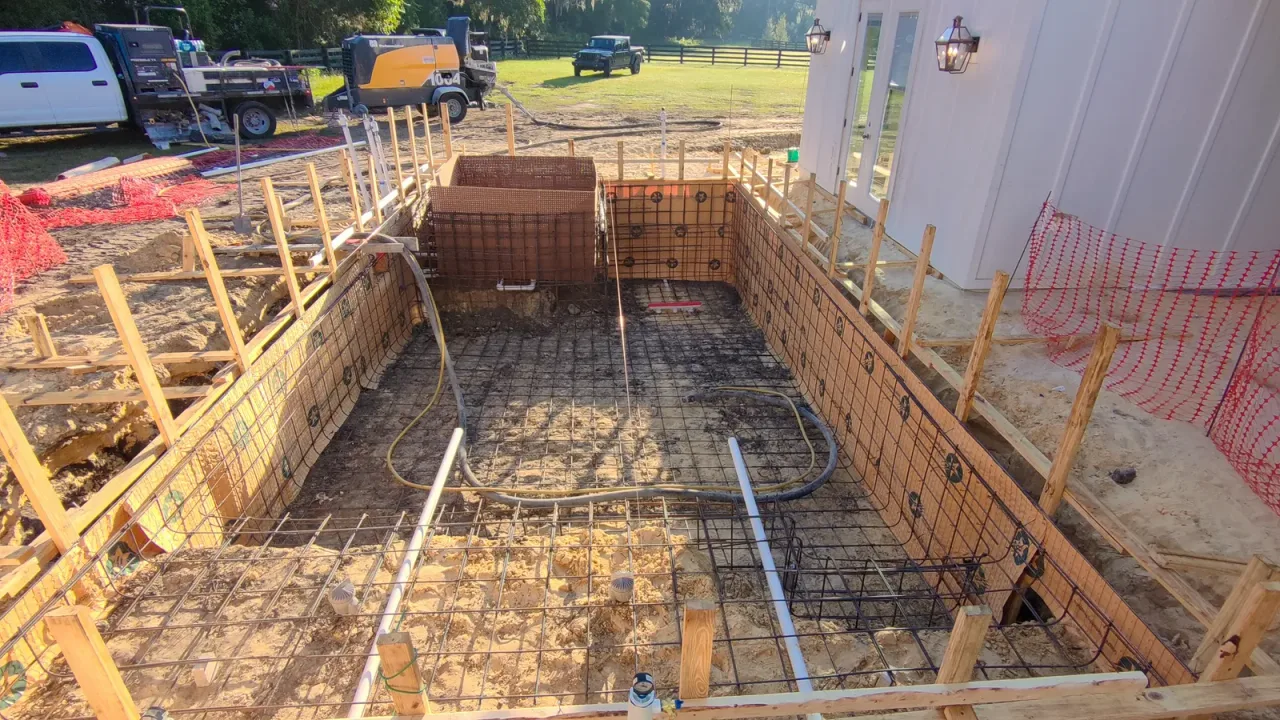 Ground view along a pool rebar cage in wood forms, with plumbing lines in place and the spa form at the far end.
