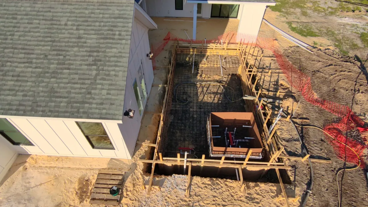 Drone view past a roof edge onto a pool rebar cage, with the spa form and its red and white plumbing lines inside it.