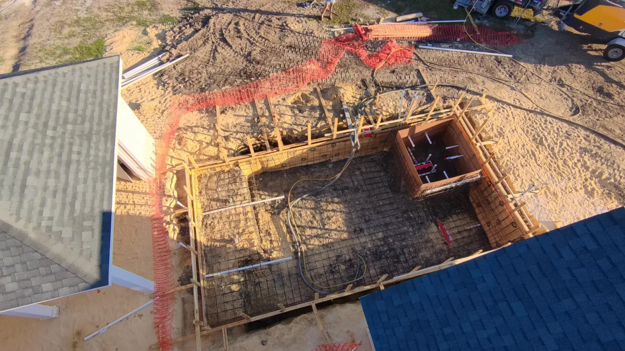 Overhead view of a pool rebar cage in wood forms, with a square spa form and plumbing stubs, between two roof edges.