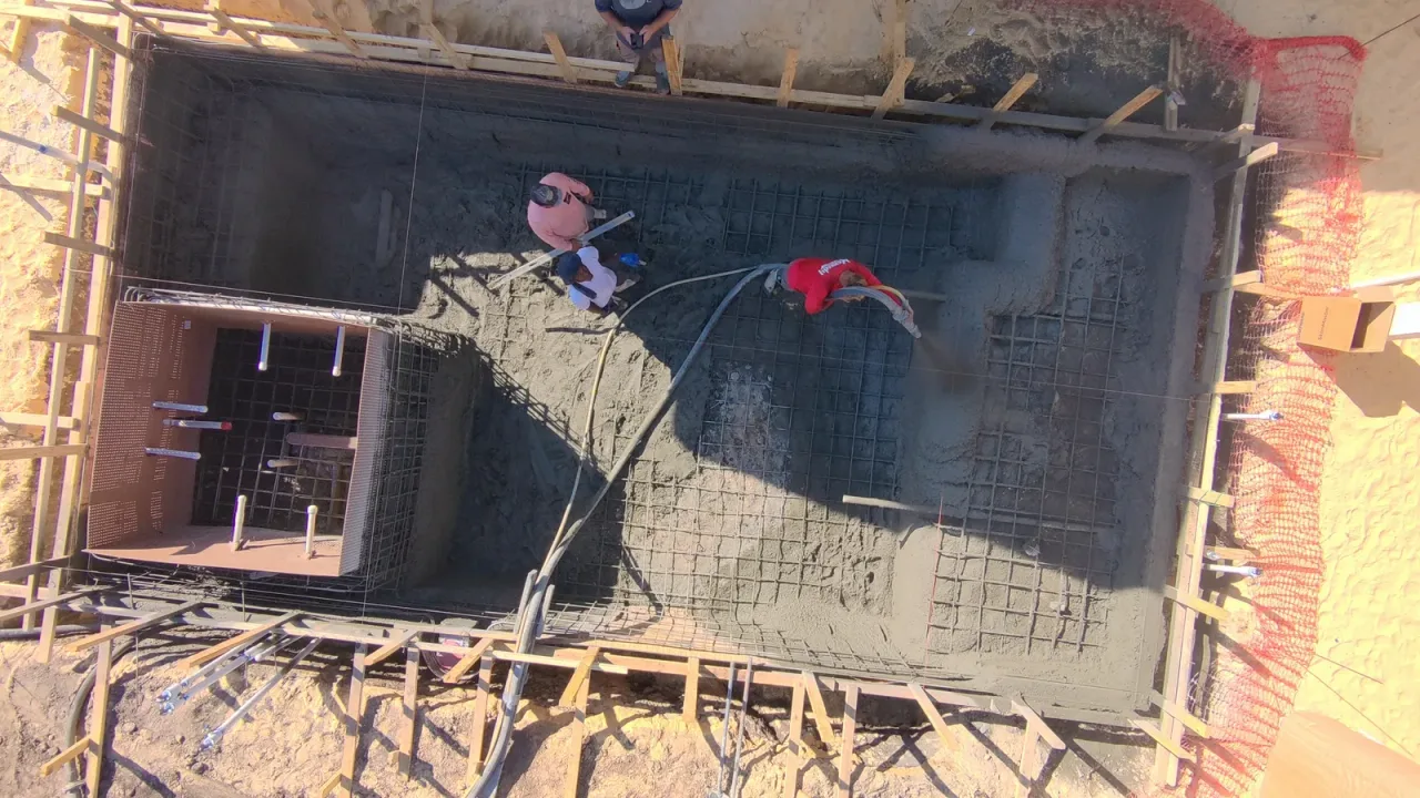 Top-down drone view of a crew shooting gunite over the rebar of a rectangular pool, with the square spa form still open.