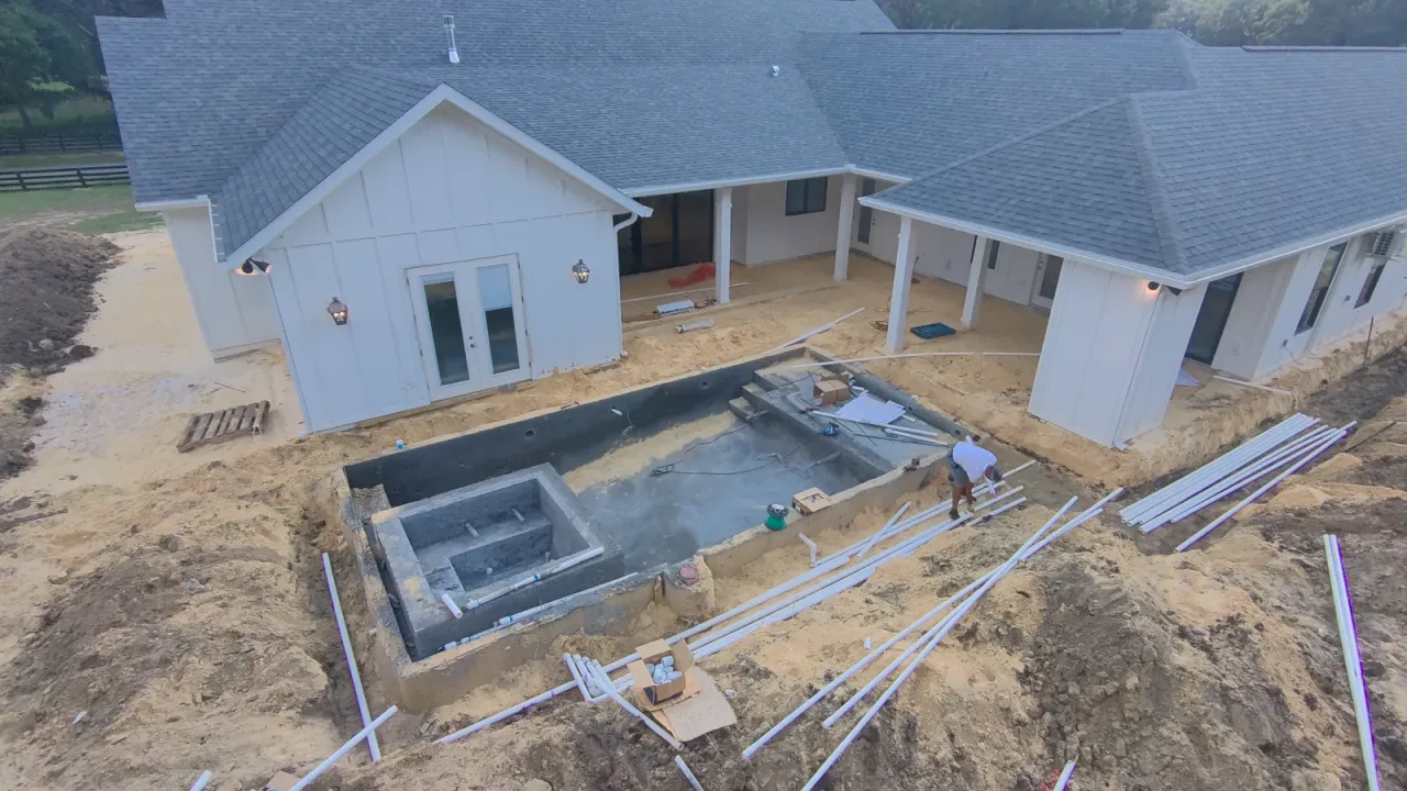 Aerial view of a gunite pool shell with its spa, and lengths of PVC pipe laid out on the sand behind a white house.