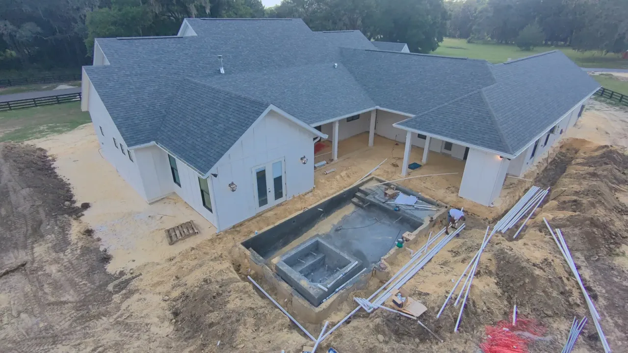 Wide aerial view of a new gunite pool shell set into the back of a large white house, with pipe laid out and trees around.