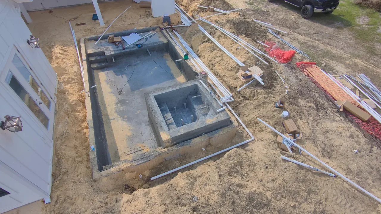 Gunite pool shell with a square spa and a trench of PVC plumbing lines running alongside it, next to the house.