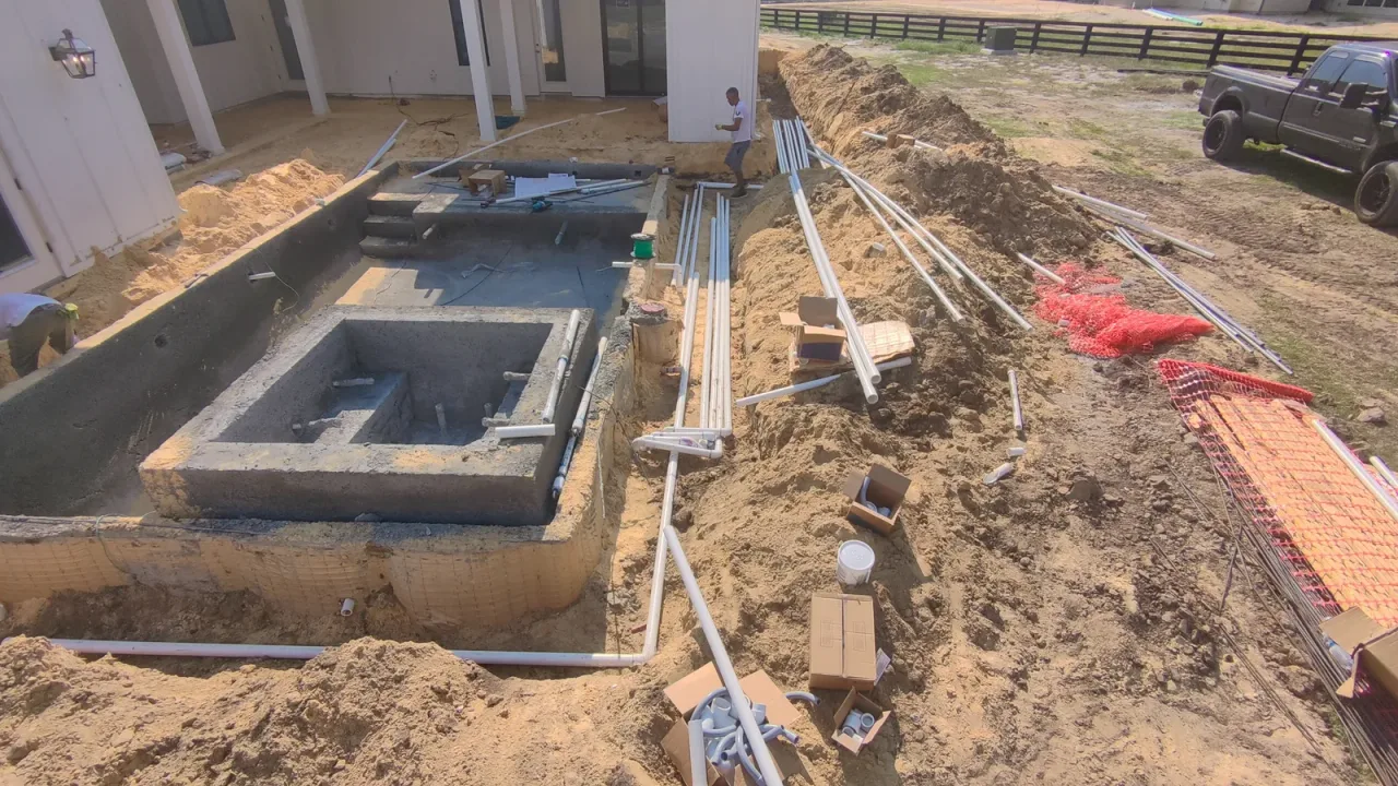 PVC plumbing lines running in a trench from a gunite spa toward the far end of the site, with fittings boxed on the sand.