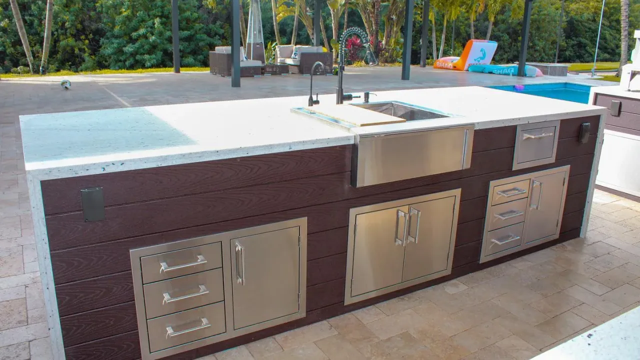 Outdoor kitchen island with a stainless farmhouse sink, drawers and doors set in brown composite cladding, a pool behind.