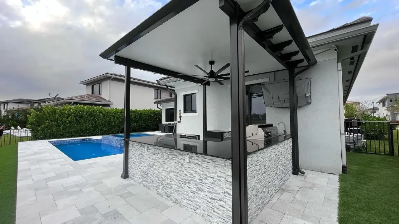 Black-framed patio cover with a ceiling fan over a stacked-stone kitchen island and grill, beside a pool and pale pavers.