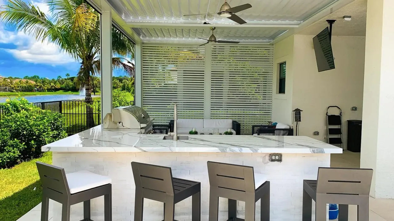 Outdoor kitchen bar with four stools facing a white veined counter with a sink and grill, under a white louvered roof.