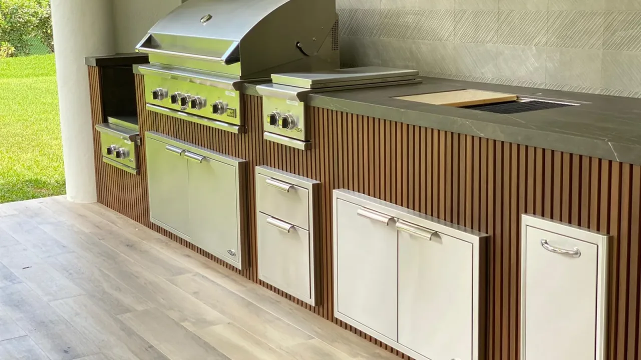 Wood-slat outdoor kitchen run with a built-in grill and side burner, stainless drawers and doors, and a dark stone counter.