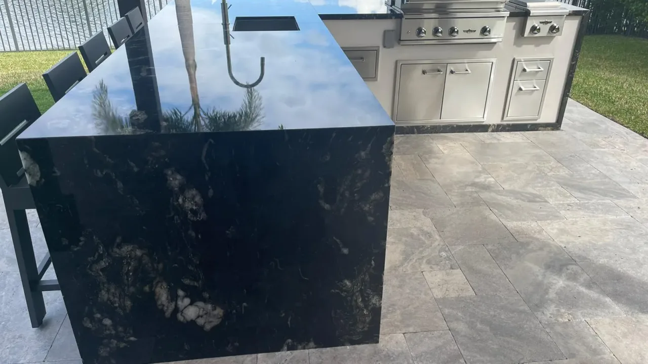 Black stone waterfall island with a sink and bar stools in front of a white kitchen run with a grill and stainless doors.