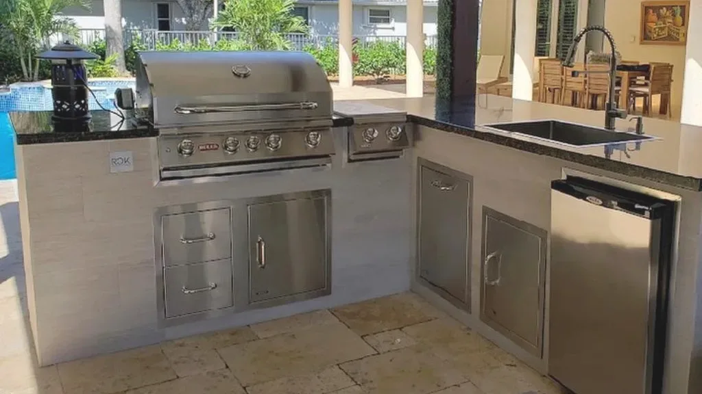 L-shaped outdoor kitchen with a built-in grill, side burner, drawers, doors, a sink and a refrigerator under a black counter.