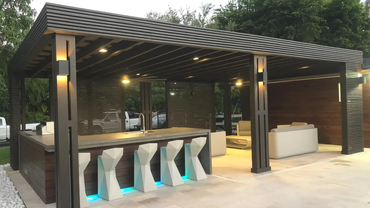 Dark slatted pavilion at dusk with lit sconces and downlights, a bar with an LED-lit base, white stools and a lounge area.