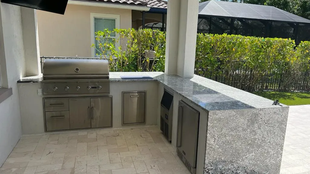 L-shaped outdoor kitchen with a white column at its corner: built-in grill, drawers, ice maker and fridge, granite top.