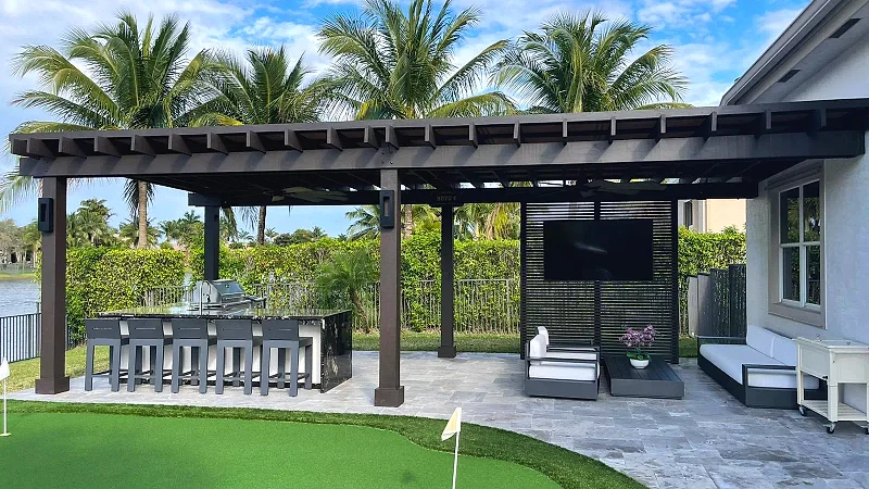 Dark pergola over an outdoor bar with stools, a TV wall and lounge seating, with a putting green and palms in front