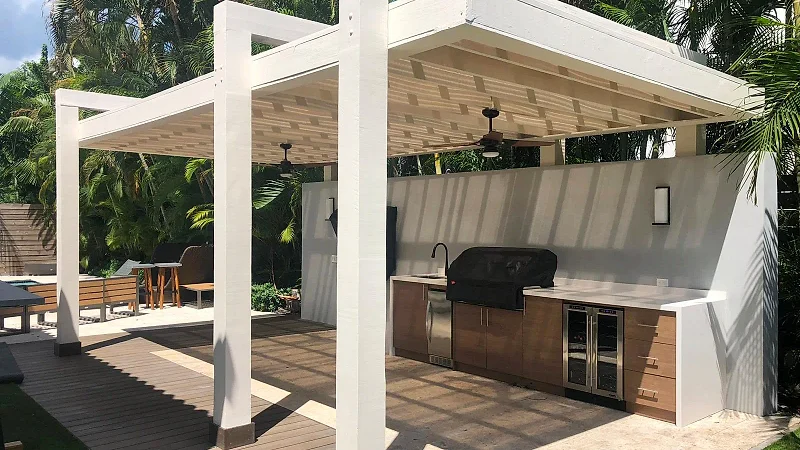 White pergola on thick square posts with ceiling fans, over an outdoor kitchen with wood-look cabinets and a covered grill