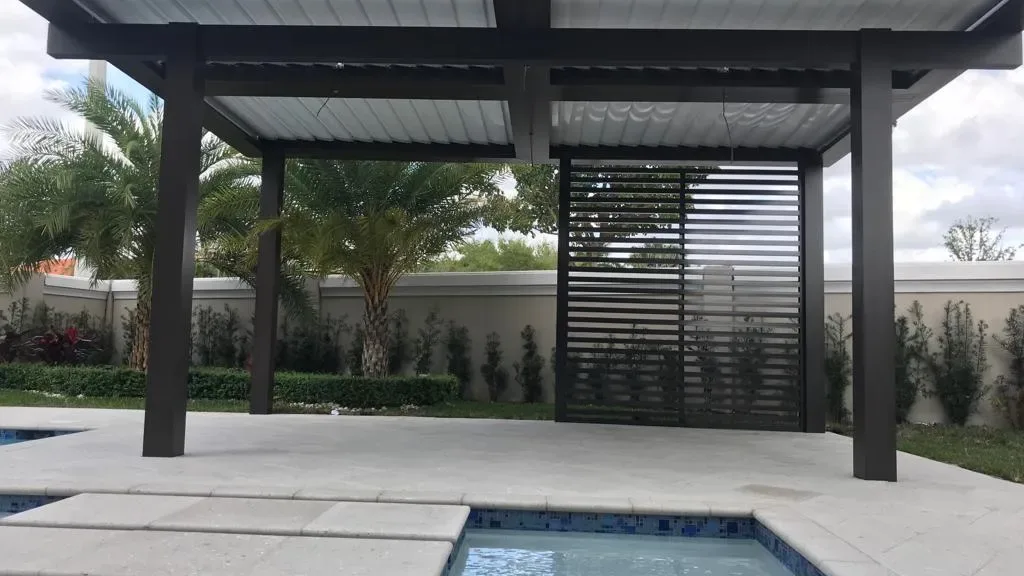 Dark aluminum patio cover with a slatted privacy screen, shading the deck at the edge of a pool with stepping pads.