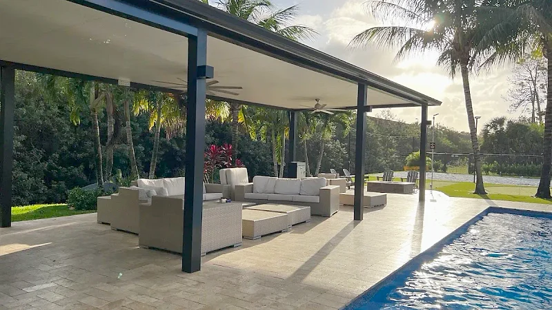 Black-framed patio cover with ceiling fans and wicker sofas on a travertine deck beside a pool, with palms backlit by the sun