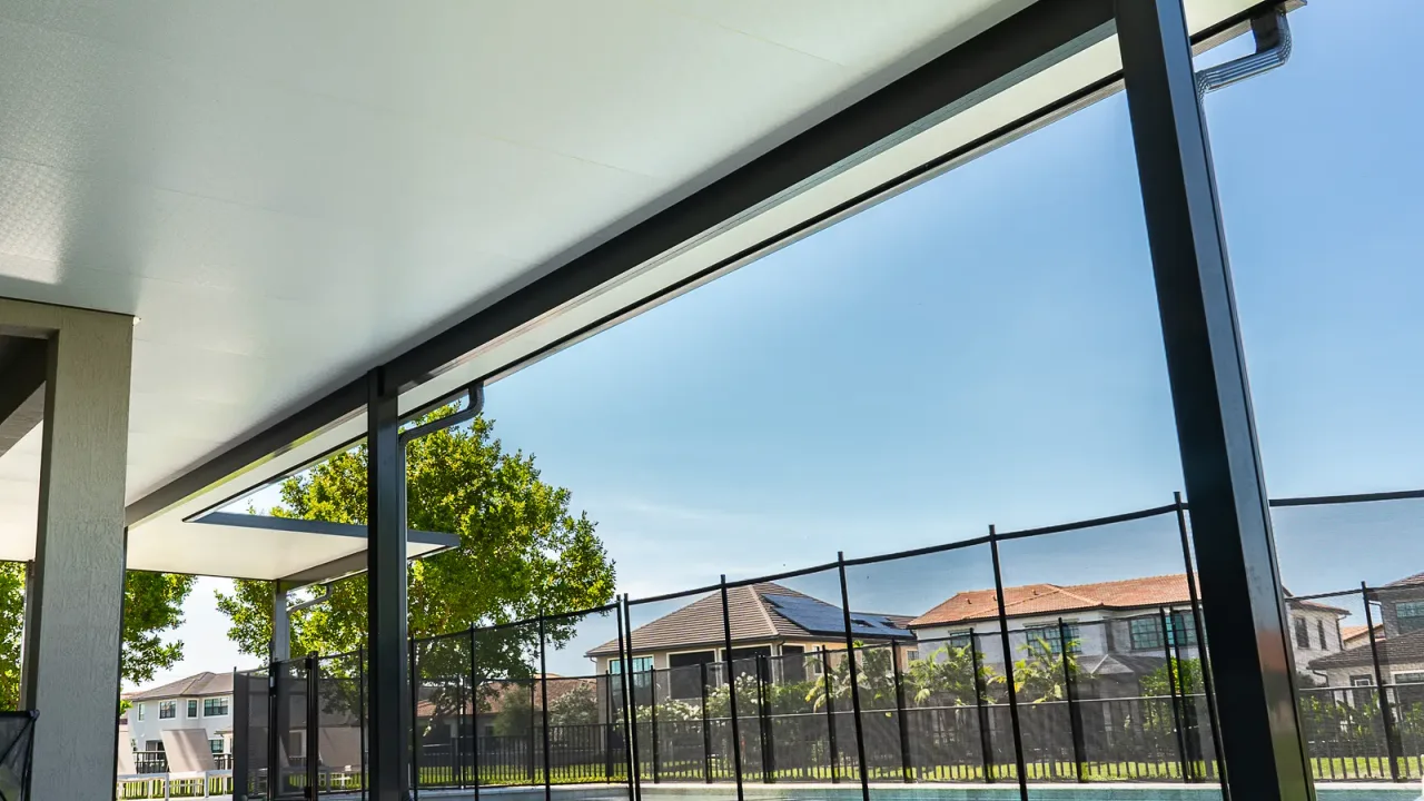 Low view under a white patio cover on black posts, a black mesh pool safety fence running just beyond them