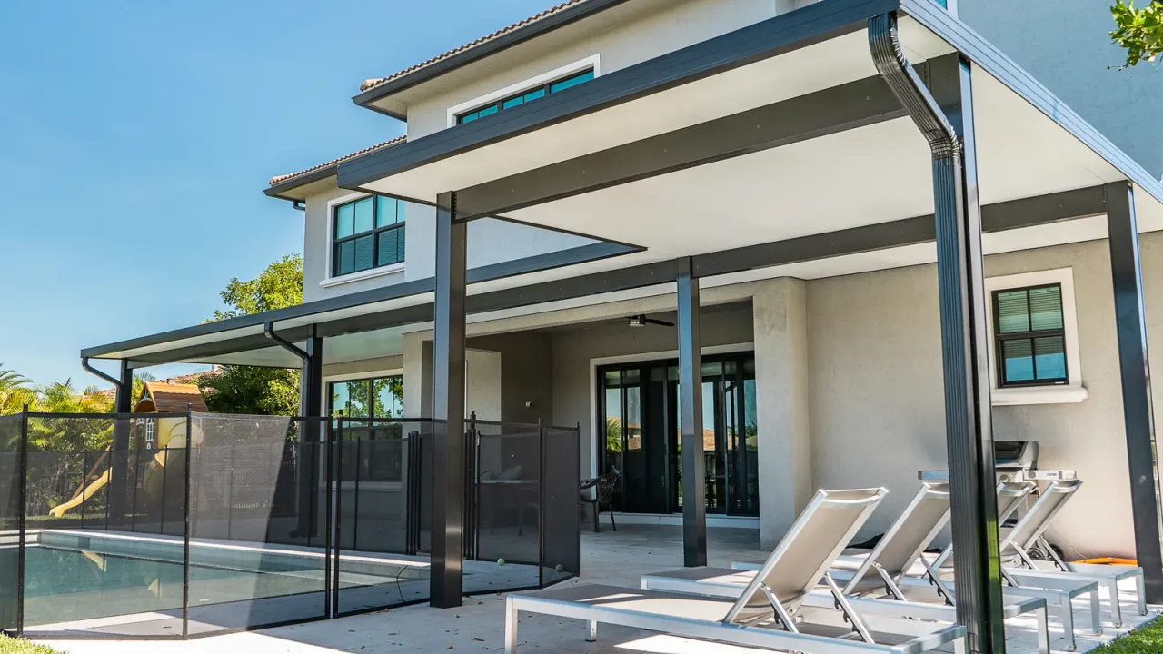 Black-framed patio cover on a two-story home beside a pool closed off by a black mesh safety fence, loungers beneath it