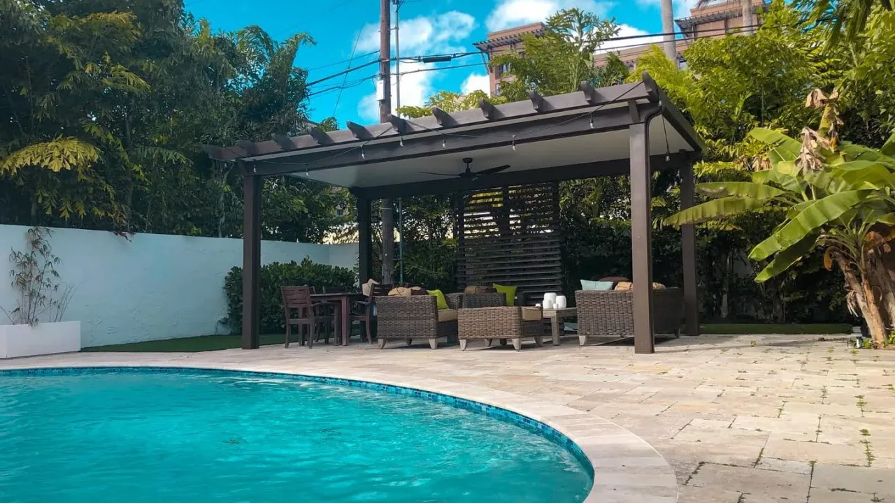 Freestanding bronze pergola with a slatted privacy screen and wicker seating on a paver deck beside a curved pool