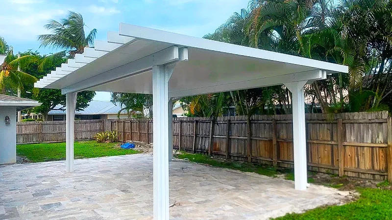 White freestanding solid-roof pergola with rafter tails over a stone paver patio, with a wood fence and palms behind