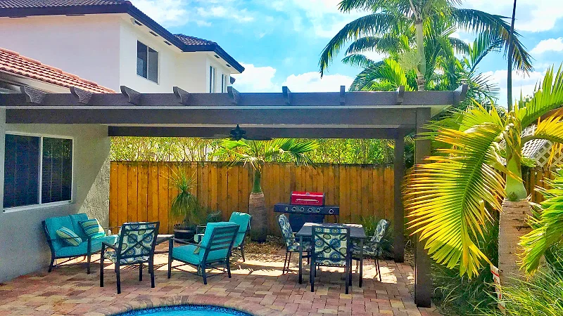 Brown pergola with rafter tails over a brick paver patio with turquoise seating, a grill and a wood fence behind