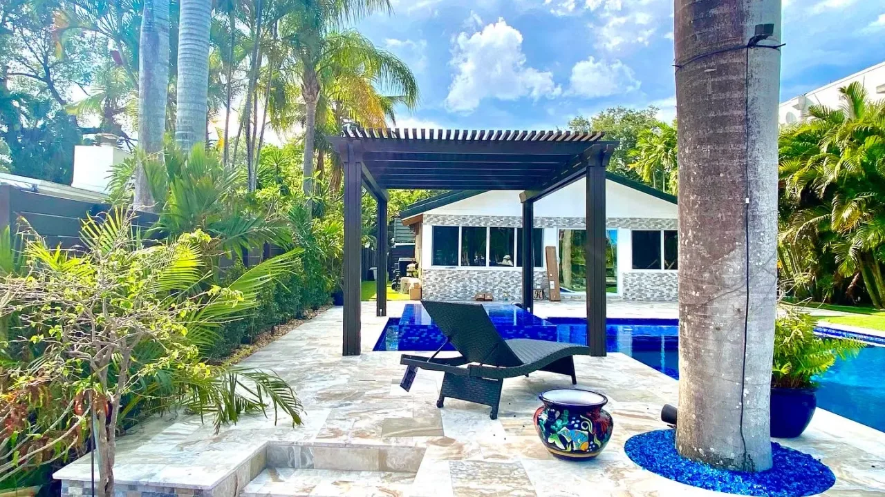 Dark bronze pergola over a blue-tiled spa beside a deep blue pool, with a wicker lounger on a stone deck.