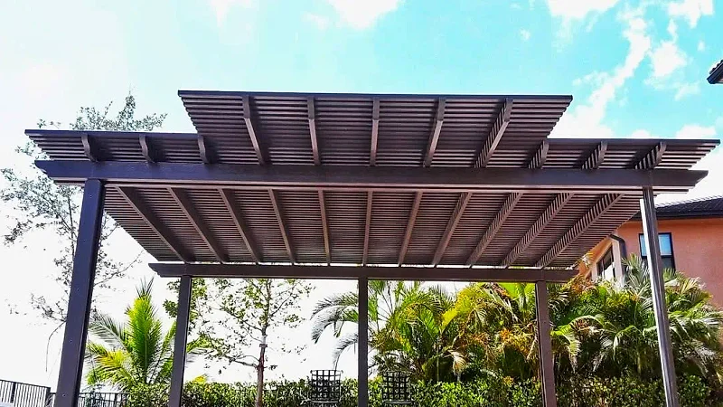 Bronze two-tier lattice pergola seen from the front against the sky, with palms, a hedge and a stucco house at right