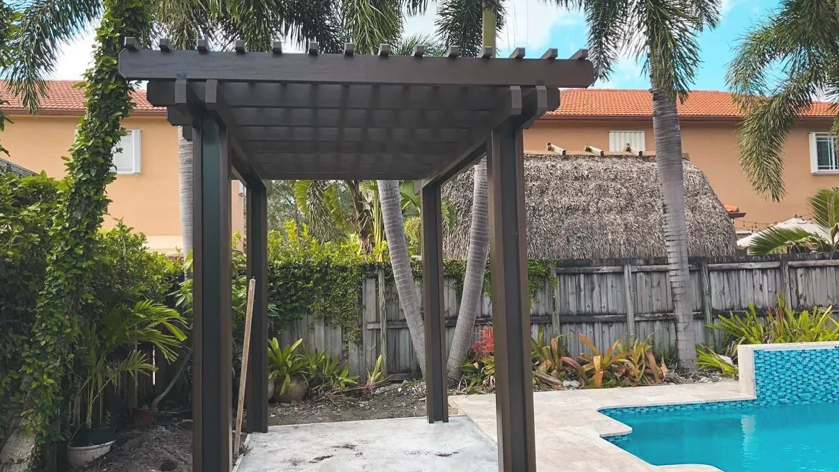 Small dark woodgrain pergola on a new concrete pad a few feet from the blue-tiled edge of a pool