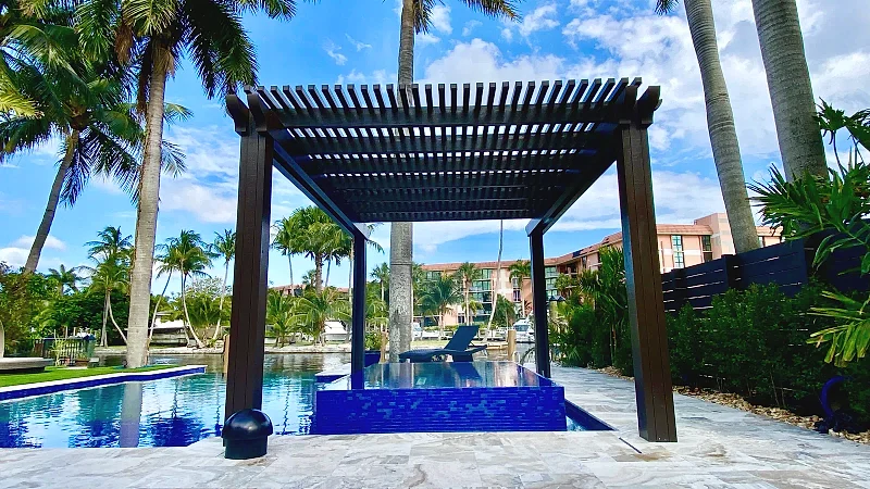 Dark freestanding lattice pergola over a raised blue-tiled spa on a stone deck, with palms and a canal behind