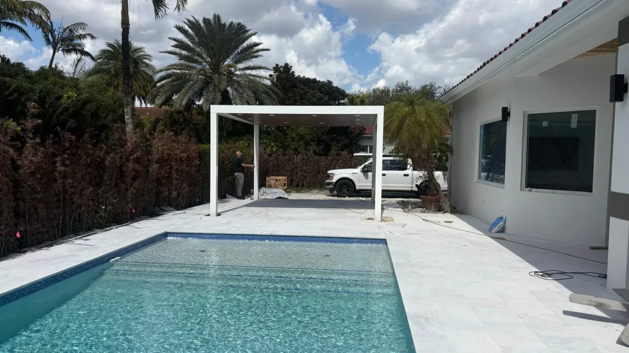 White freestanding flat-roof pergola going up at the end of a new pool, beside a white house with a pale stone deck.