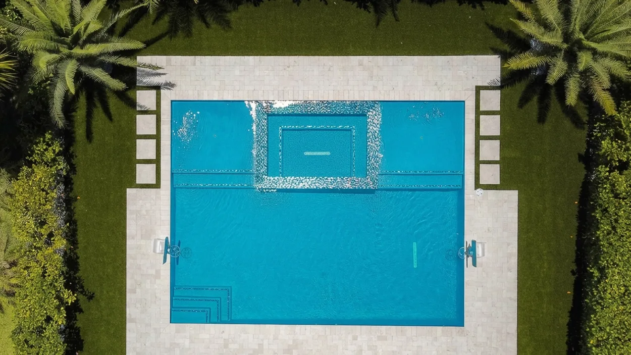 Overhead view of a rectangular pool with a square spa set into it, water spilling over the spa rim on all four sides in a band of white foam, on a travertine deck with handrails at the pool edges and a lounger and blue umbrella at the bottom of the frame.