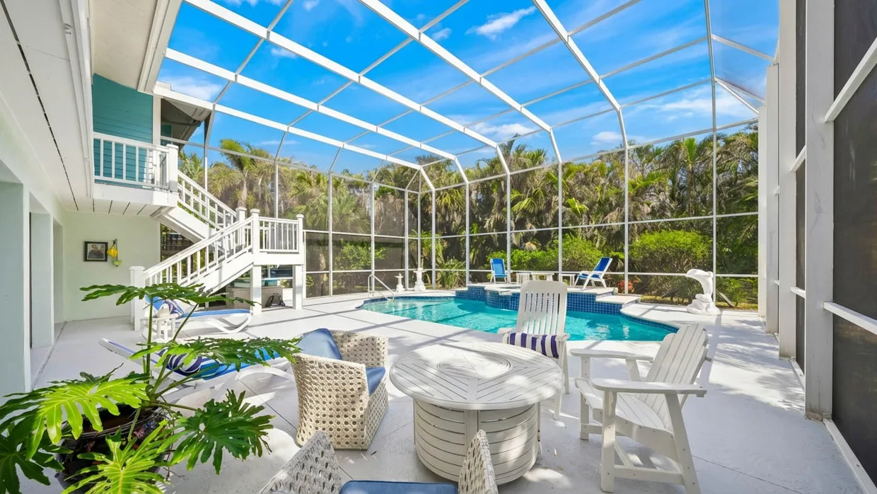 Curved pool and raised spa under a white-framed screen enclosure, with white Adirondack chairs around a round table on a painted concrete deck and an exterior staircase climbing to the teal-sided upper floor of the house.