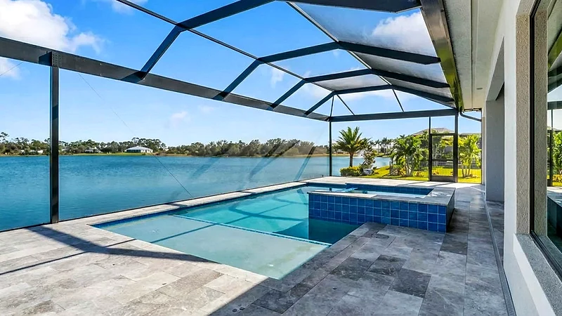 Pool with a raised spa finished in blue tile inside a screen enclosure, on a gray travertine deck, the frame braced with thin diagonal cables and a lake filling the view behind.