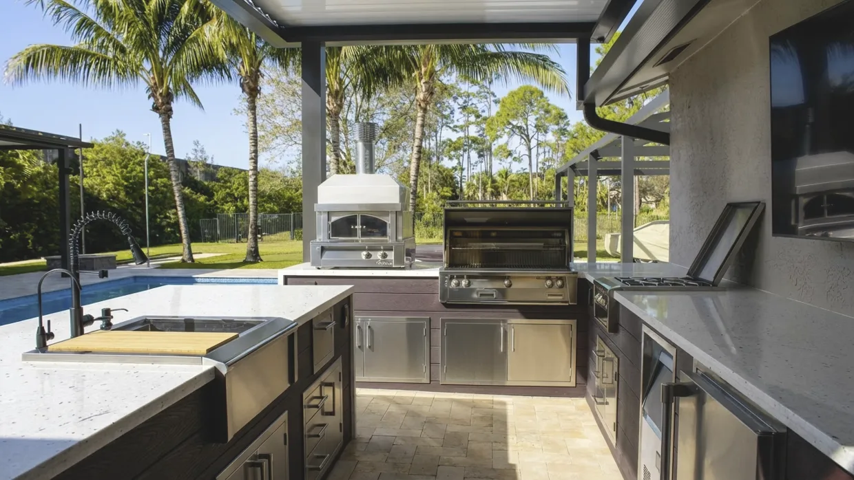 Poolside outdoor kitchen with a wood-fired oven and its chimney flue standing next to a built-in grill and a side burner, and a sink with a cutting board on the island in front.