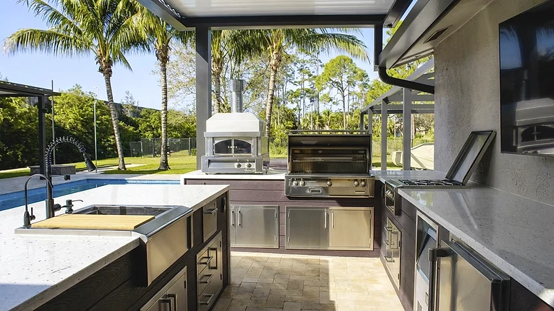 Outdoor kitchen with a sink and cutting board, a pizza oven, built-in grill and side burner, looking out to a pool and palms