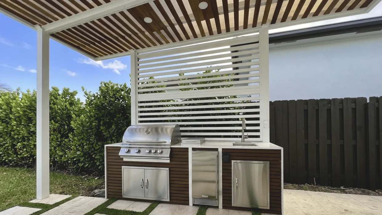 Straight-run outdoor kitchen with dark wood-slat cabinet fronts, a stainless grill, a small sink and a white counter, under a white pergola with wood slats and recessed downlights, with a white louvered privacy screen behind it and grass strips between the pavers.