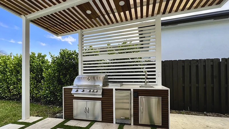 Grill island with wood-look fronts, a built-in grill, sink and refrigerator under a slatted wood pergola with a white screen