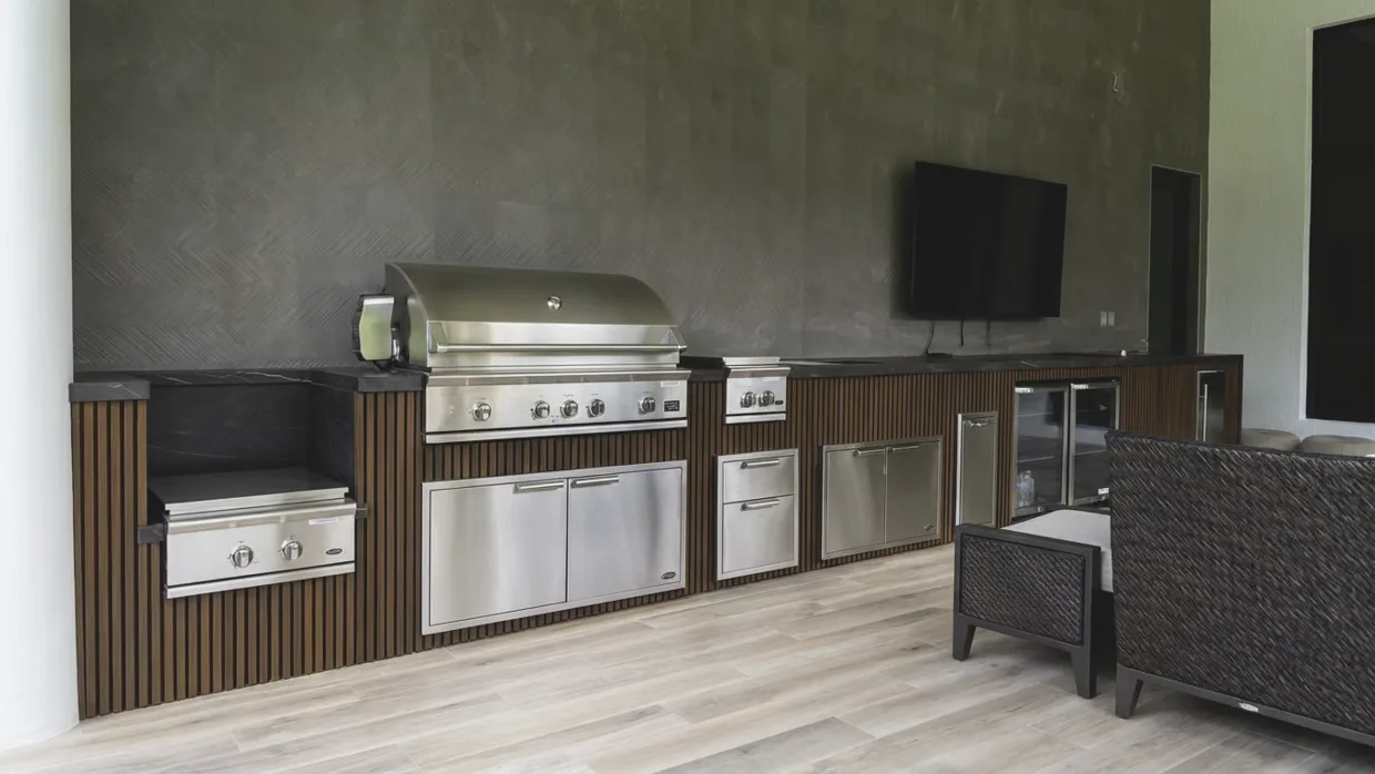 Long single-wall outdoor kitchen run against a dark textured wall: side burner, built-in grill, drawers, glass-front beverage refrigerator and a wall-mounted television above the counter.