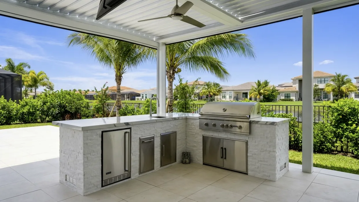 L-shaped outdoor kitchen clad in white stacked stone under a white louvered roof with a ceiling fan, with a built-in stainless grill and stainless doors and drawers, and a pond with tile-roofed houses behind.