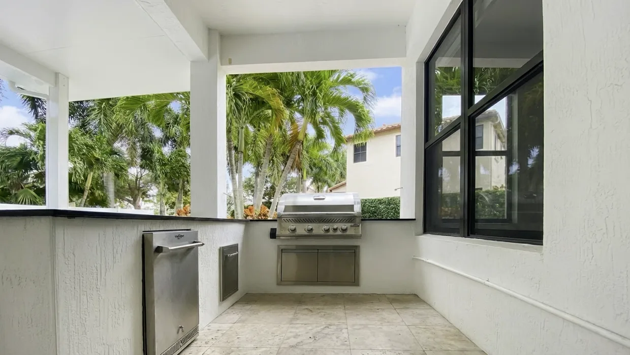 Covered lanai outdoor kitchen: a built-in stainless grill and a small stainless access door set into a white stucco base under a dark countertop, an under-counter stainless unit at the end of the run, and a travertine floor.
