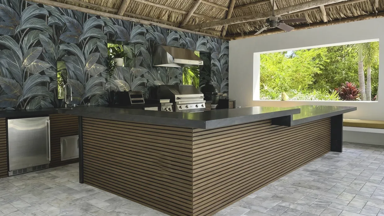 Outdoor kitchen under a thatched chickee roof with slatted wood cabinet fronts, a dark countertop wrapping into a bar, a stainless hood over the grill and a sink at the far left.