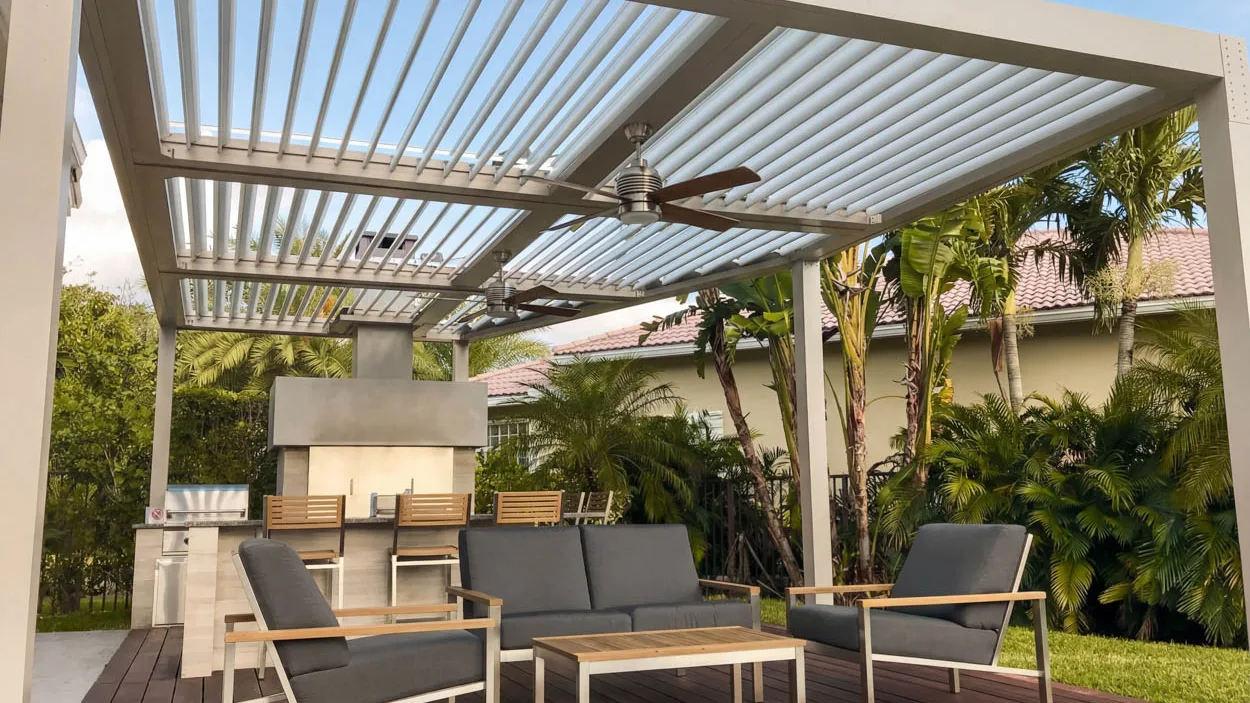 Cream louvered roof over a wood deck, with two ceiling fans hanging under it, a stainless steel built-in grill in a masonry counter and a tall outdoor fireplace at the far end.